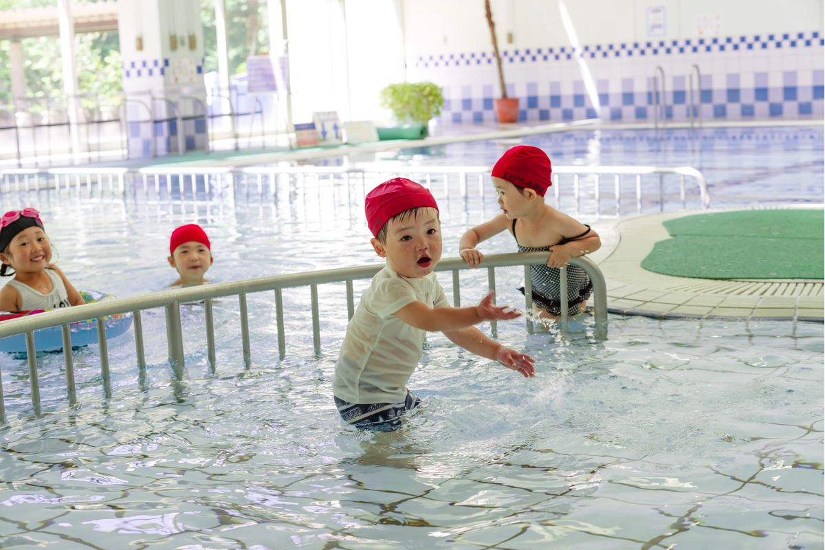 屋内の浅いプールで、手すりにつかまって立つ小さな子どもたち