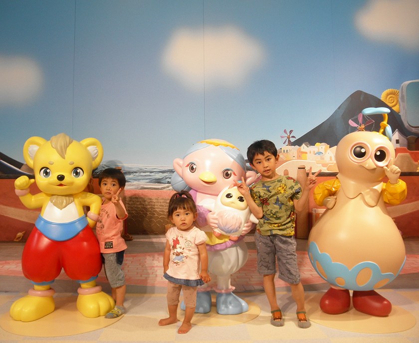 屋内の展示で、裸足の小さな子どもがキャラクターの人形と並んでいる