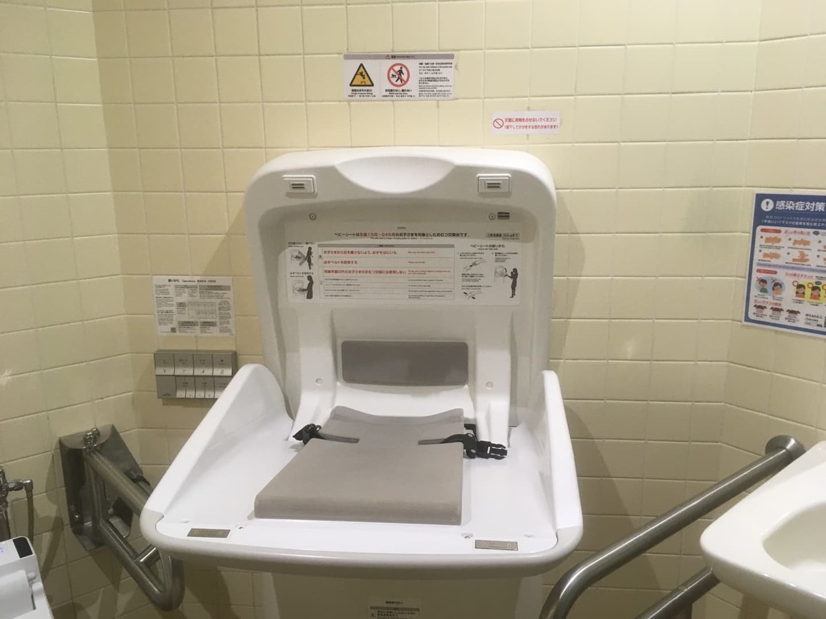 淡いタイル張りのトイレの壁に、折りたたみ式の白いおむつ交換台が開いた状態で取り付けられている
