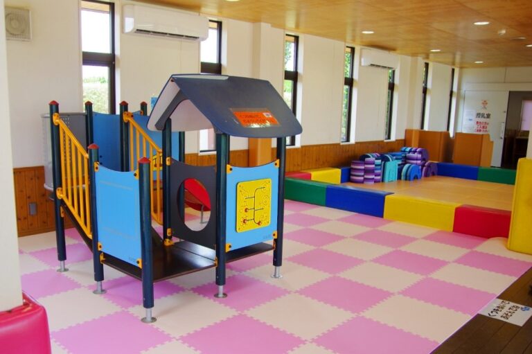 室内のプレイルーム。マットを敷いた床にすべり台つきの遊具が置かれ、奥にブロックのコーナーと授乳室の入口がある