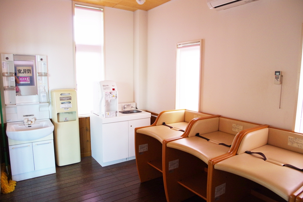 おむつ替えシートが3台並ぶ部屋。奥に洗面台とおむつ用のゴミ箱、給湯器が置かれている