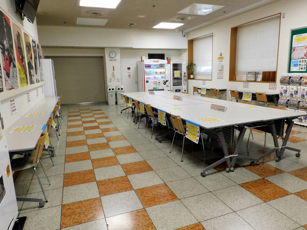 長机と椅子が並んだ部屋。奥に飲み物の自動販売機、右手にカプセルトイの機械が置かれている