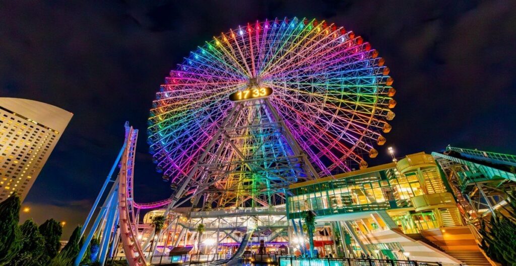 夜に虹色にライトアップされた大観覧車。手前にジェットコースターのレールと建物の明かりが見える