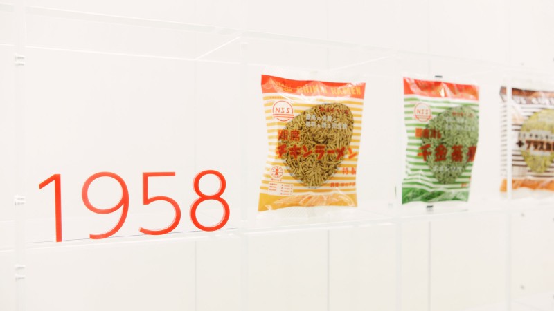 白い壁に「1958」と赤い数字が入り、その横に初期のインスタントラーメンの袋が透明のケースで並んでいる