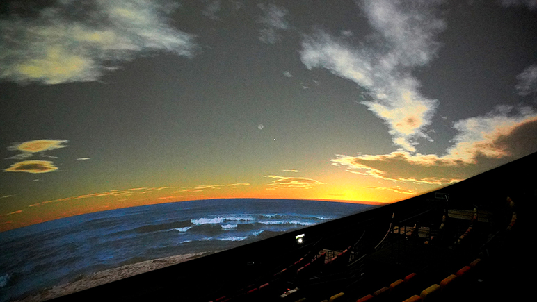 ドーム一面に映る夕暮れの海と空。手前に客席の列が見える