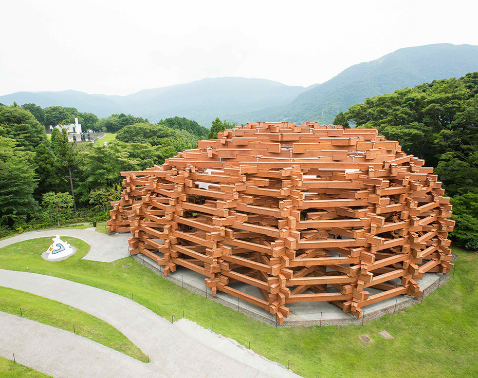 芝生の中に建つ、木の角材を積み上げた大きなドーム型の建物。奥に箱根の山なみが見える