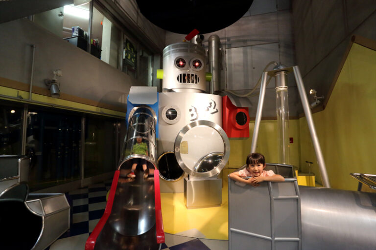 銀色の大きなロボットの形をした遊具。胴体から金属のすべり台と筒状のトンネルが伸び、中から小さな子どもが顔を出している