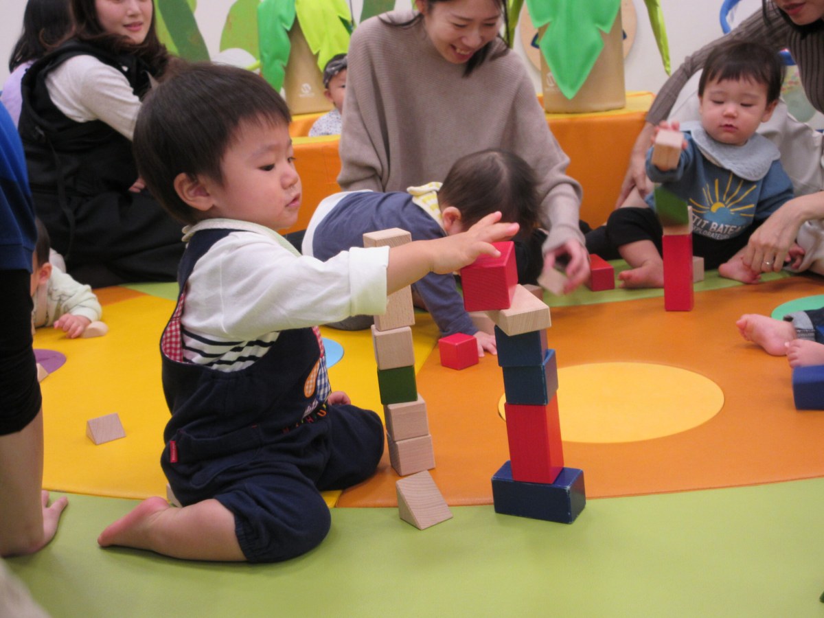 床に座って積み木を高く積み上げる子どもと、周りで見ている親子