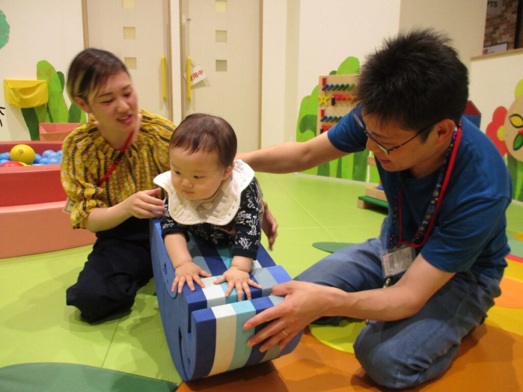 青い筒状のやわらかい遊具に手をつく赤ちゃんを、プレイリーダーと大人が両側から支えている
