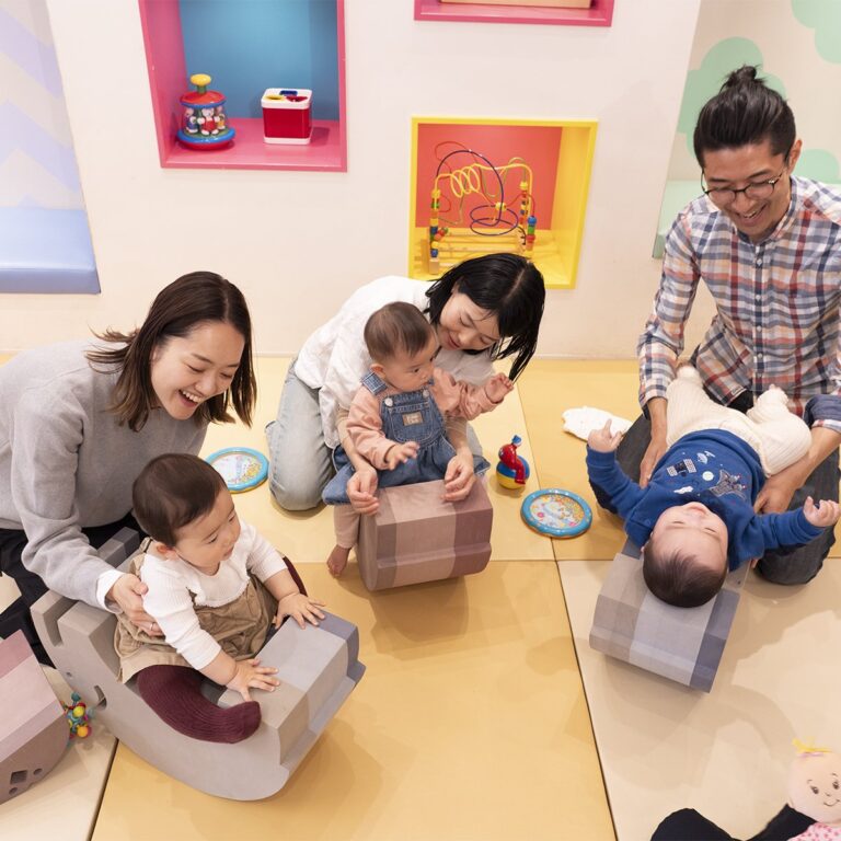 やわらかいブロックにまたがったり寝転んだりする赤ちゃん3人と、そばで支える大人たち