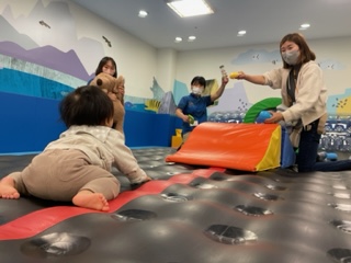 赤いマットの上でハイハイする赤ちゃんと、色つきの布を持って前で待つスタッフ