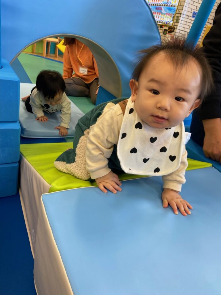 やわらかいマットの段に手をついてハイハイする赤ちゃんと、奥のトンネル型遊具でハイハイするもう1人の赤ちゃん