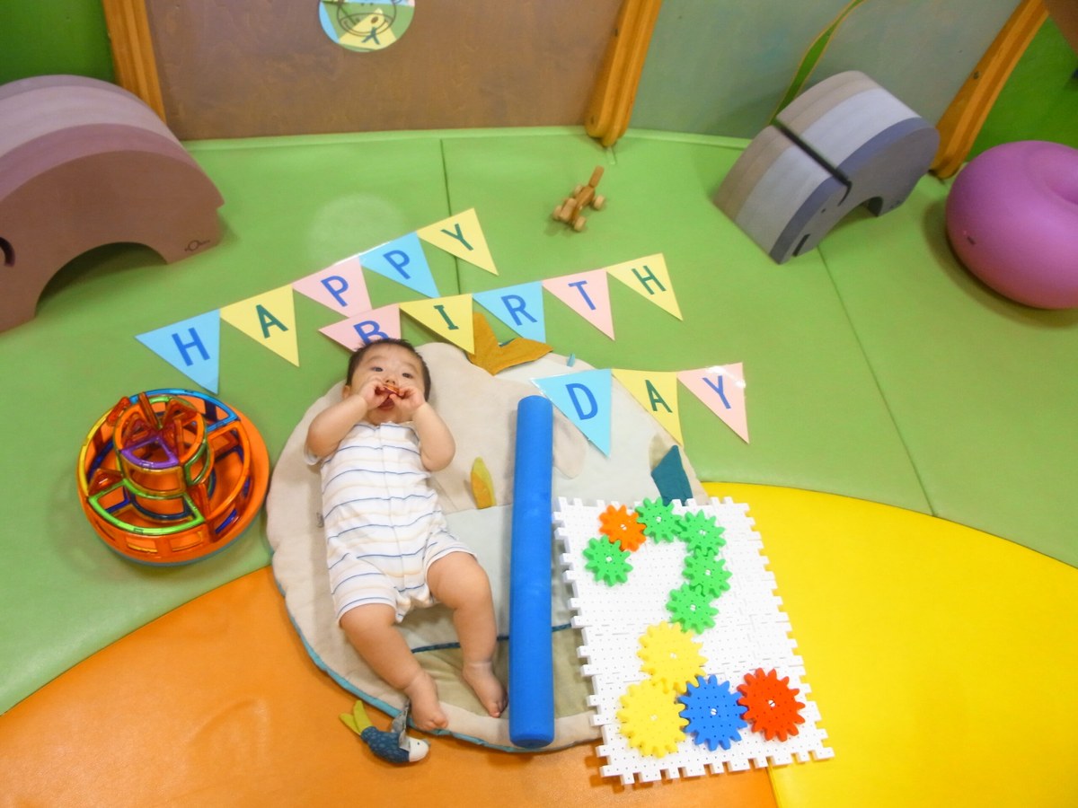 やわらかい床のベビーガーデンで、HAPPY BIRTHDAYの旗を背にして寝転ぶ赤ちゃん