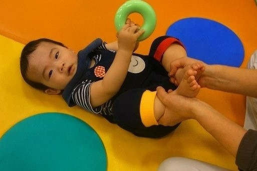 黄色いマットの上であおむけになり、輪の形のおもちゃを握る赤ちゃんと、足を支える大人の手