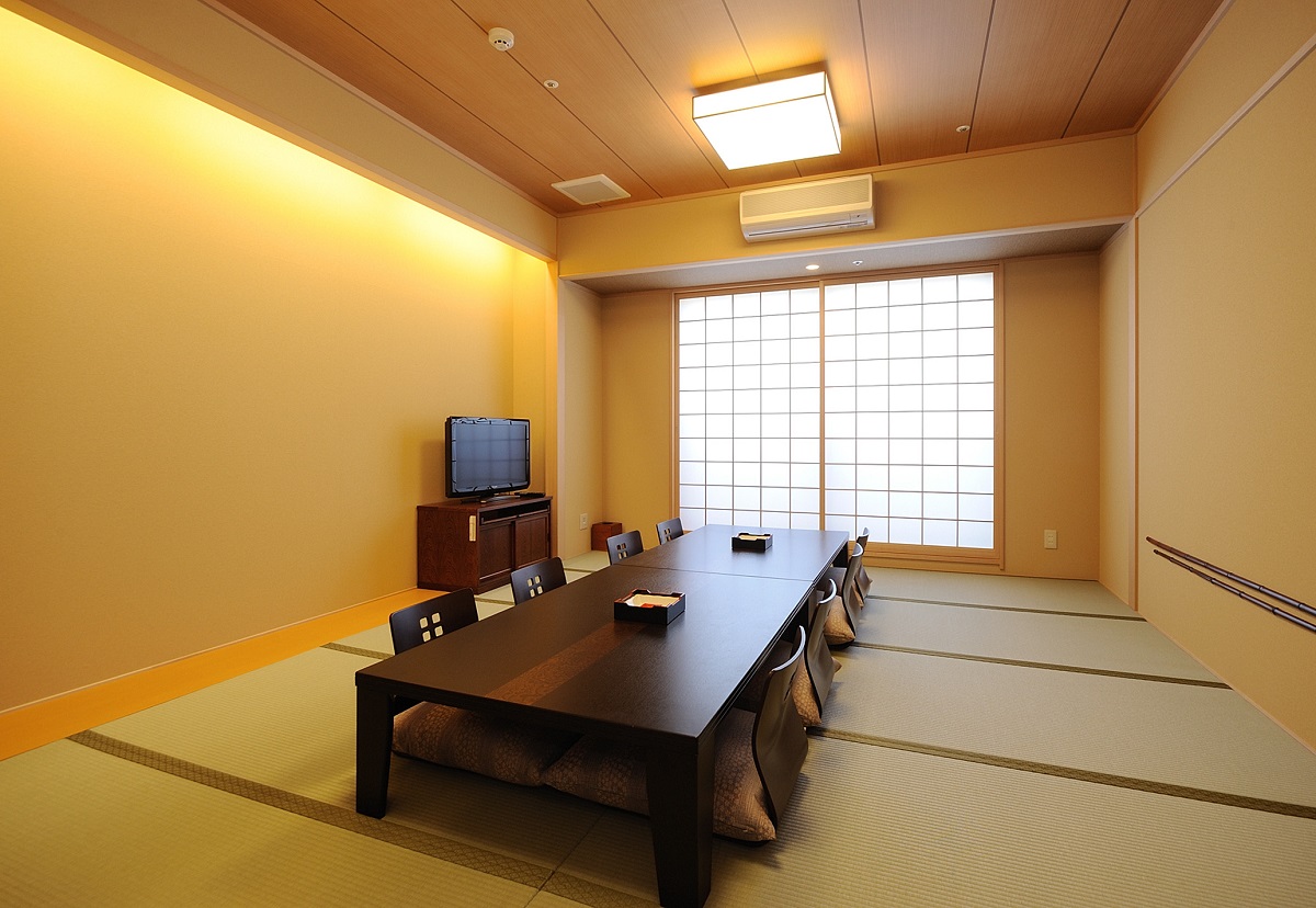 貸部屋。畳敷きの和室に黒い座卓と座椅子が並び、障子の窓から光が入る。奥にテレビ