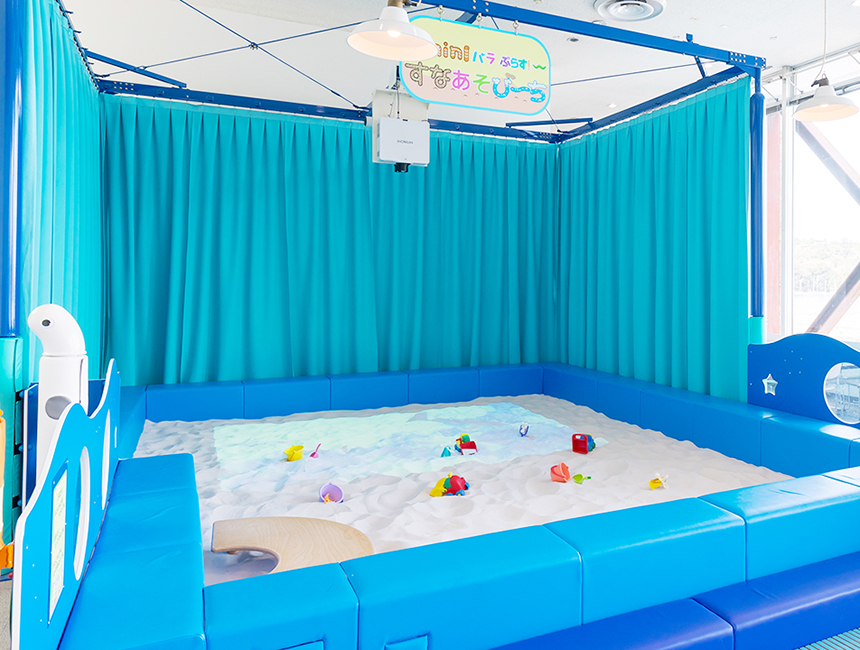 水色のカーテンに囲まれた屋内の砂場。白い砂の上にスコップやバケツが置かれている