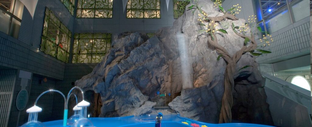吹き抜けの屋内にそびえる高さ10メートルの岩山。細い滝が落ちていて、足もとに青い浅いプールが広がっている