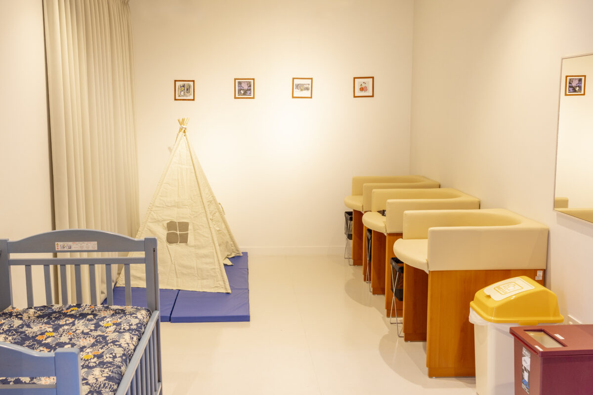 白い部屋におむつ交換台が4台並び、左手にベビーベッド、奥に子ども用の三角テントが置かれている