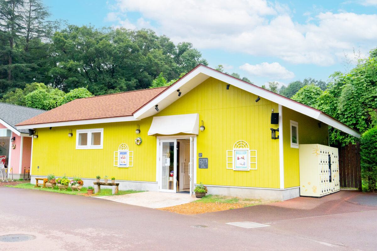 黄色い板壁に赤茶色の屋根の平屋。白い庇の下に出入口があり、前に花のプランターが並ぶ