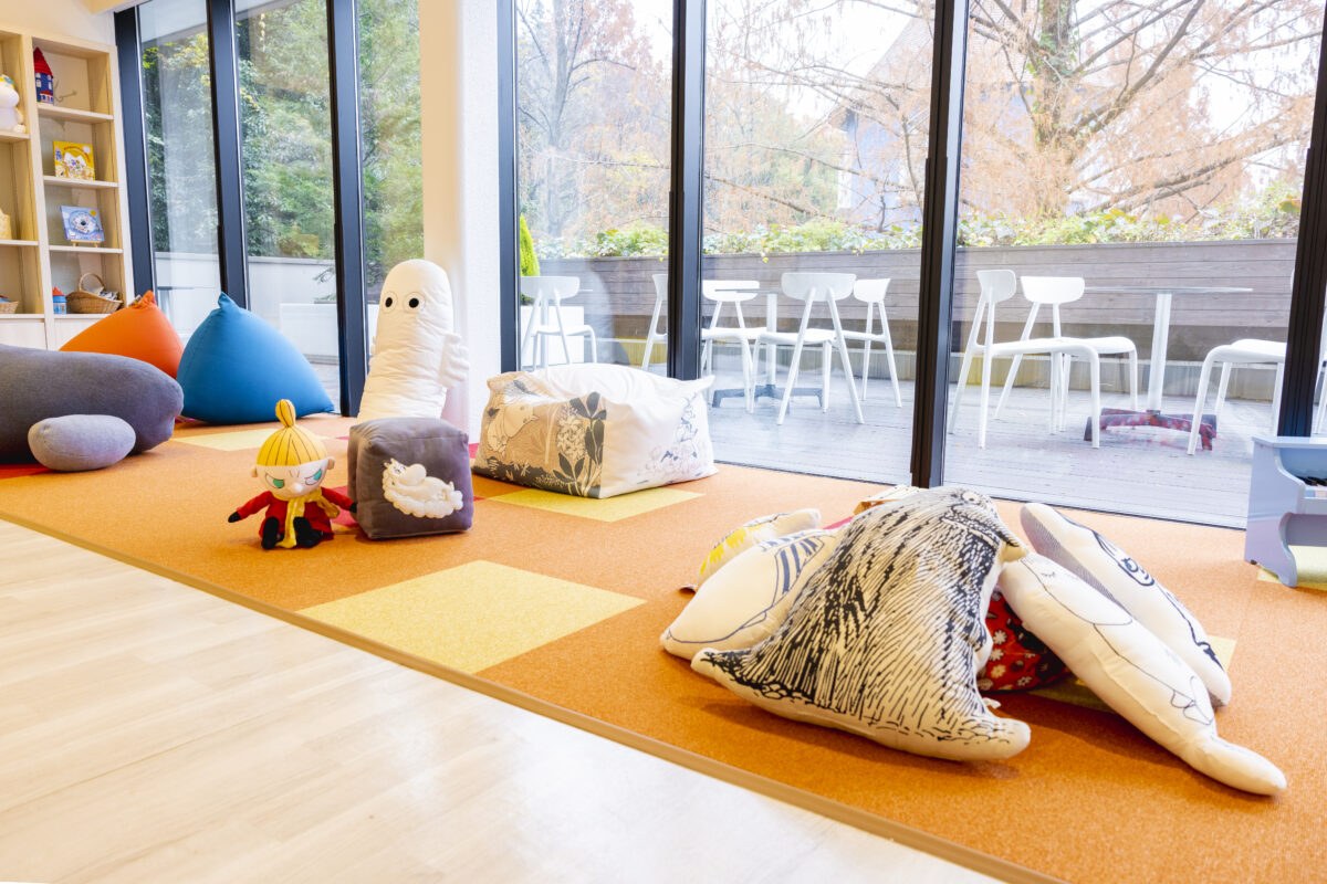 オレンジ色のカーペットにクッションとぬいぐるみが置かれた部屋。奥は一面のガラス窓で、外にテラス席が見える