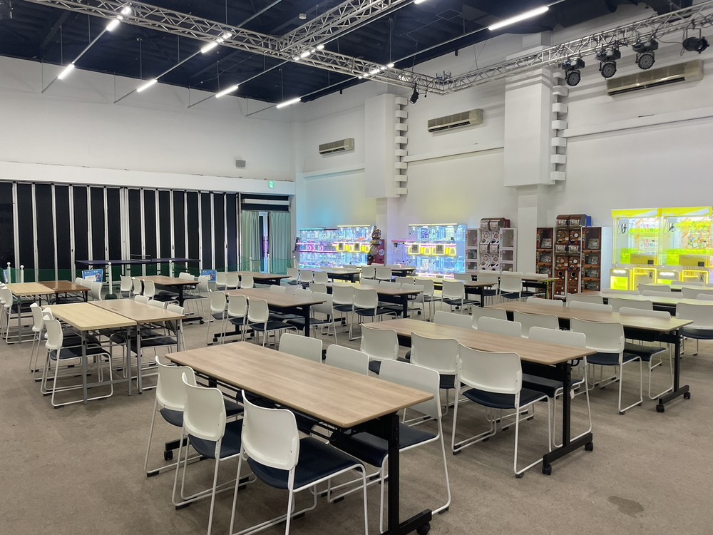 白い壁の広い屋内スペースに、木目のテーブルと椅子が何列も並ぶ。奥にクレーンゲームの機械が置かれている