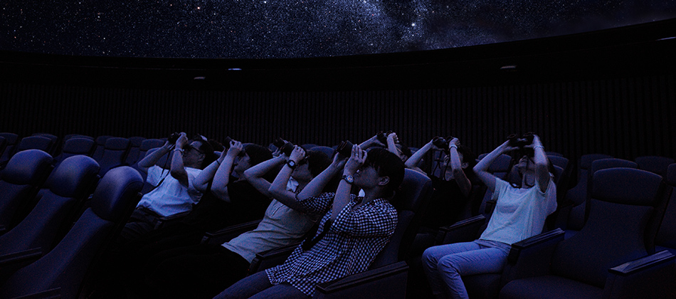 暗いドームの下、座席に座った人たちが双眼鏡で星空を見上げている