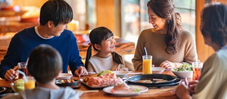 一枚板のテーブルを囲んで、大人3人と子ども2人が笑っている。中央に生肉の盛り合わせと黒い焼き網が置かれている