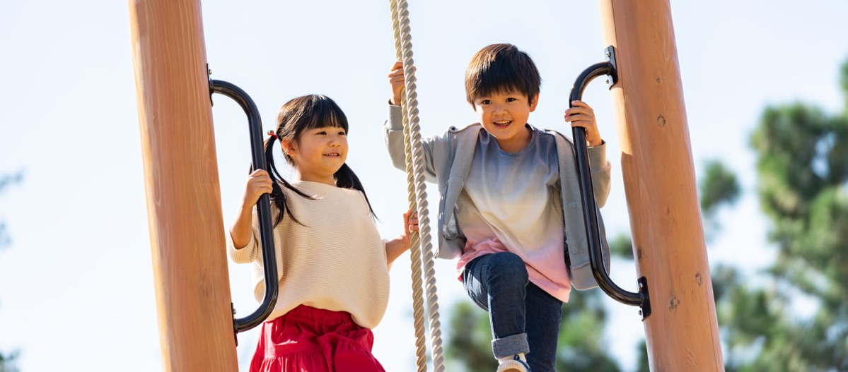 青空の下、木の柱にはさまれた遊具の上で、女の子と男の子が黒い手すりとロープをつかんで立っている