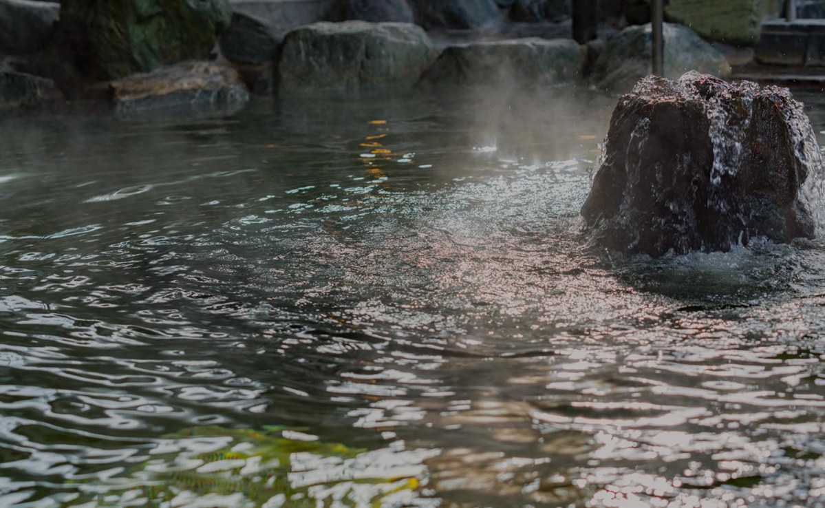 岩に囲まれた露天風呂の湯面から湯気が立ちのぼり、右手の岩から湯が流れ落ちている