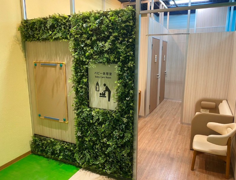 緑の植栽で囲まれた入口に「ベビー休憩室 Baby Care Room」の表示。奥に個室の扉が並び、手前にひとりがけの椅子が置かれている