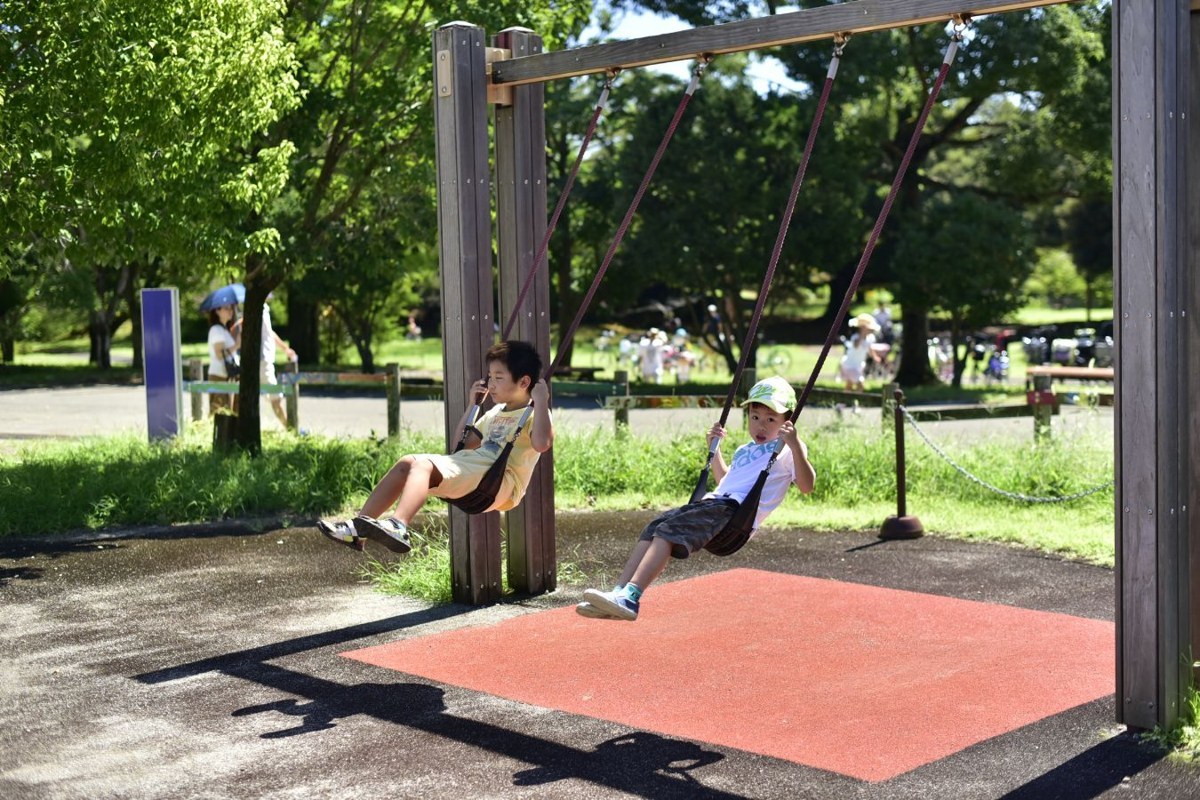 木製の枠のブランコに子ども2人が乗って遊んでいる。ブランコの下は赤いゴム舗装になっている
