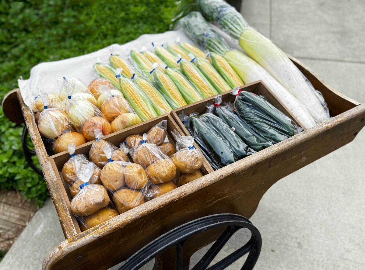 木の手押し車に並べられた野菜。とうもろこし、じゃがいも、葉物が袋に入って売られている
