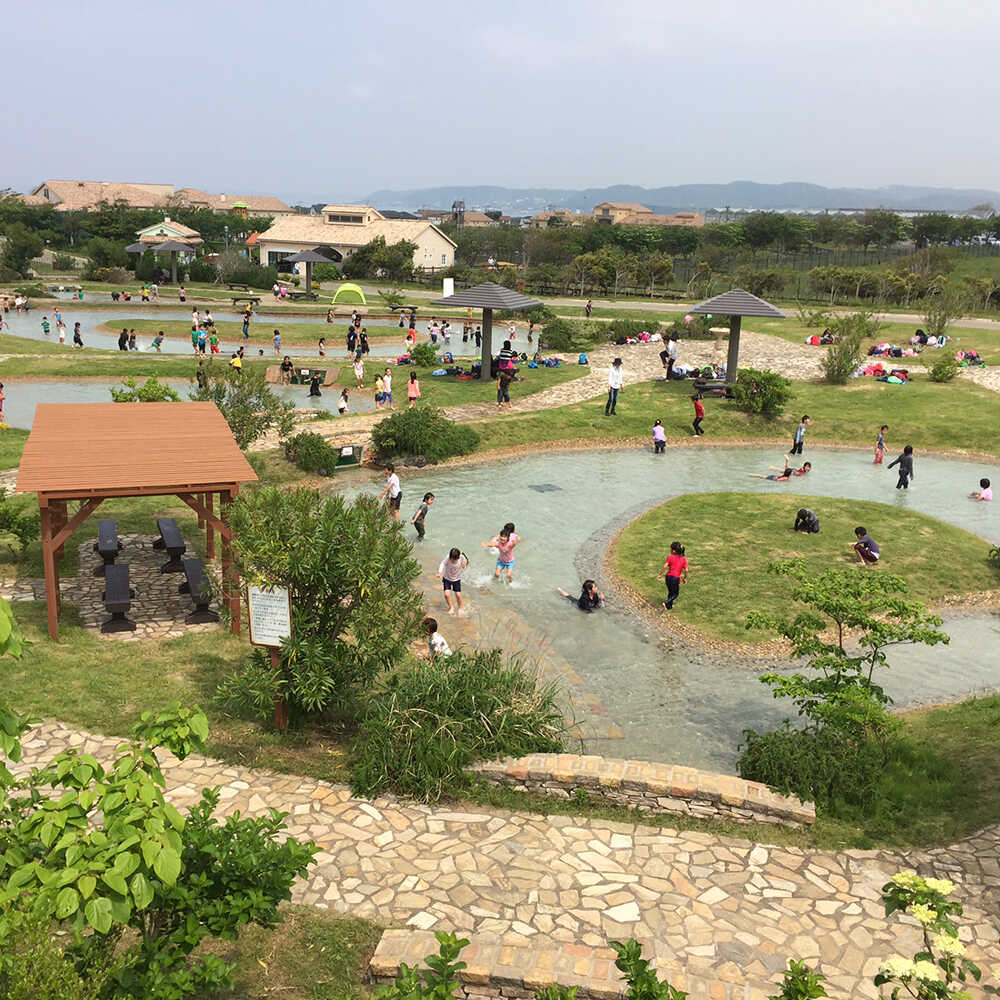 浅い水路のような池で子どもたちが水遊びをしている。まわりは石畳と芝生で、東屋が建っている