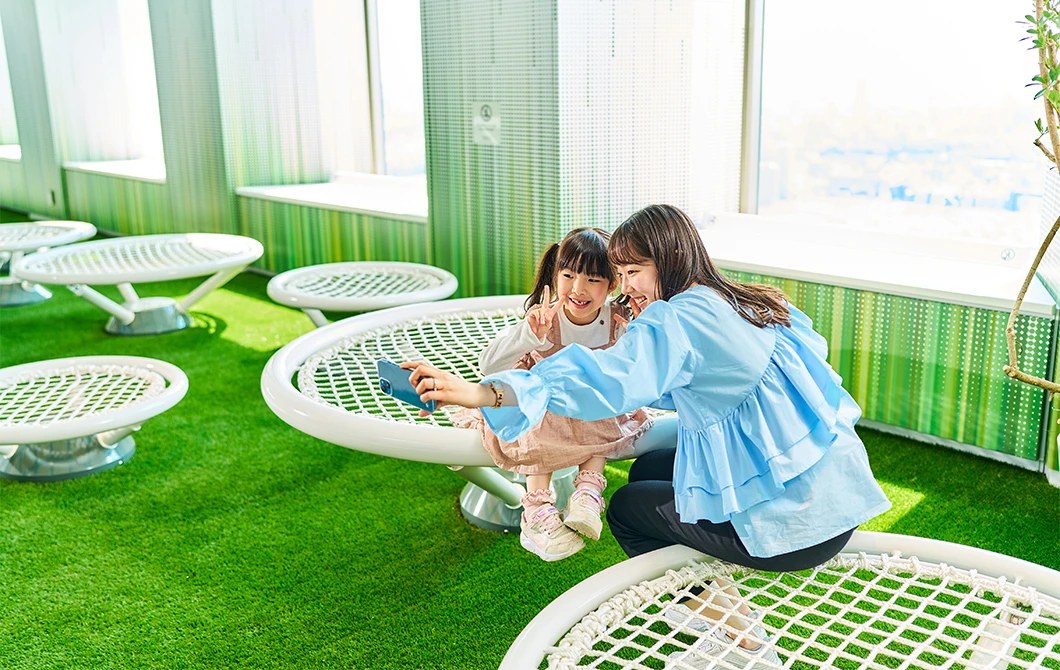 人工芝の上に白い網状の丸いベンチがいくつも並び、その上で子どもと大人が座っている。奥は窓で外の景色が見える