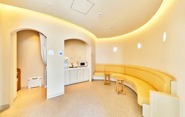 曲線を描くベンチとテーブルが置かれた明るい部屋。壁際に給湯器と電子レンジのカウンターがあり、左手にアーチ型の入口の個室が並ぶ