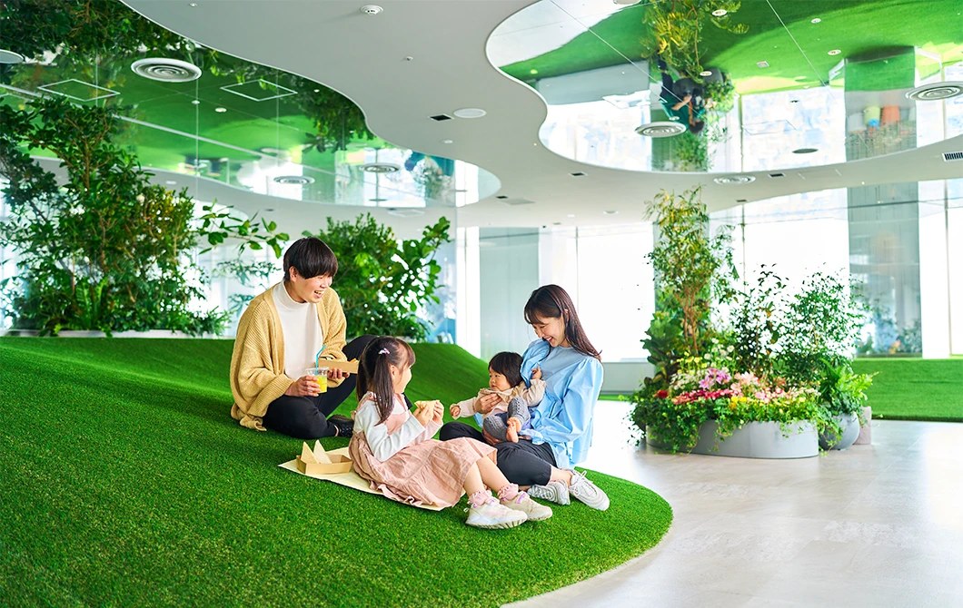 室内の人工芝でできたゆるやかな丘の上に家族が座り、紙袋を広げて食べている。まわりに植栽と大きな窓がある