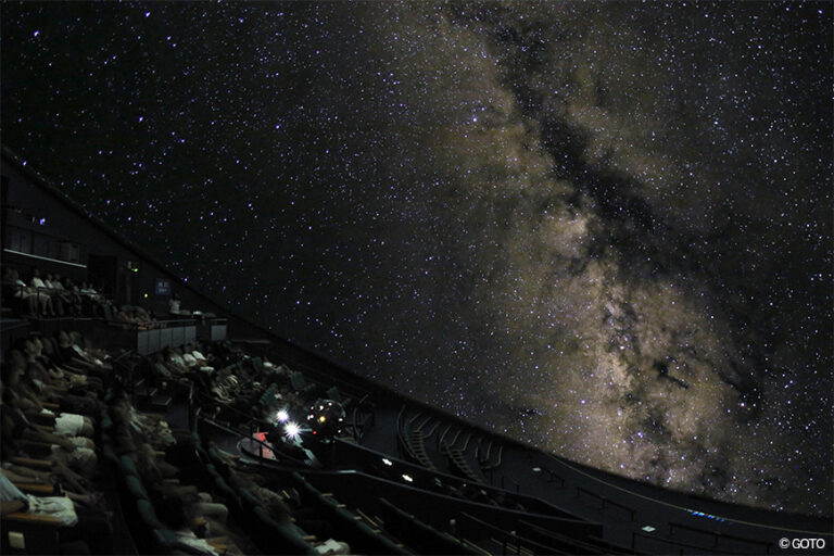 ドーム一面に天の川が広がる星空。手前に傾斜した客席のシルエットが並んでいる