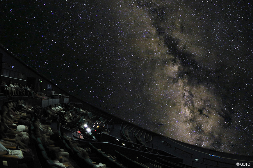 ドーム一面に天の川が広がる星空。手前に傾斜した客席のシルエットが並んでいる