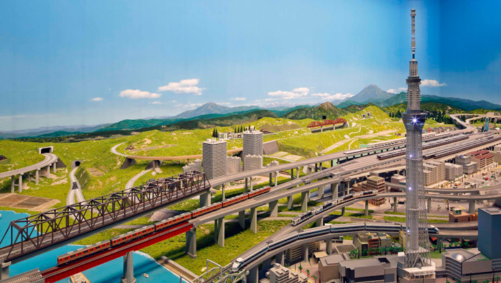 山と川を模した巨大なジオラマ。高架橋の上を模型電車が走り、右奥にスカイツリーの模型が立っている