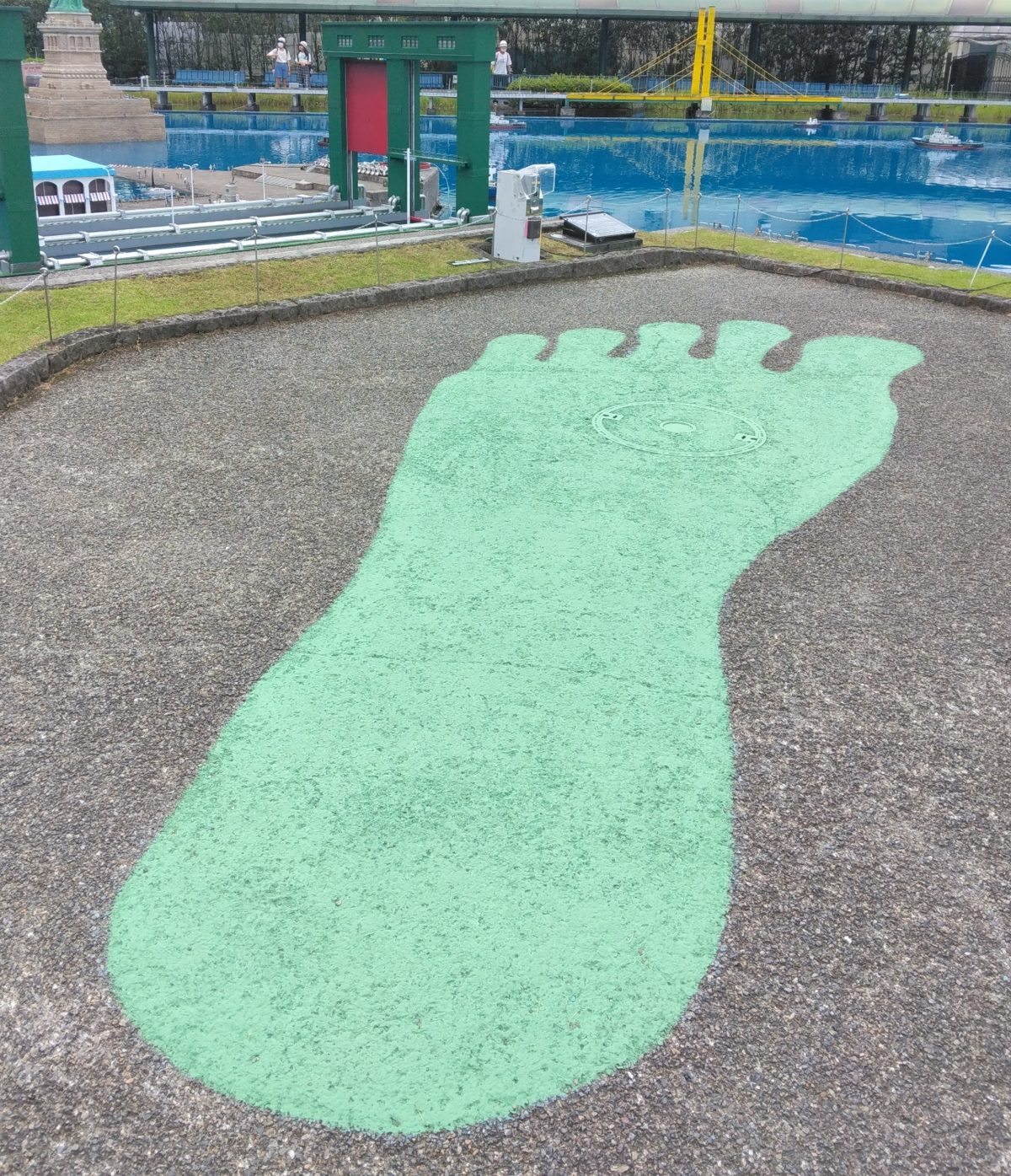 地面に描かれた大きな緑色の足あと。奥に青い水面とミニチュアの建物が見える