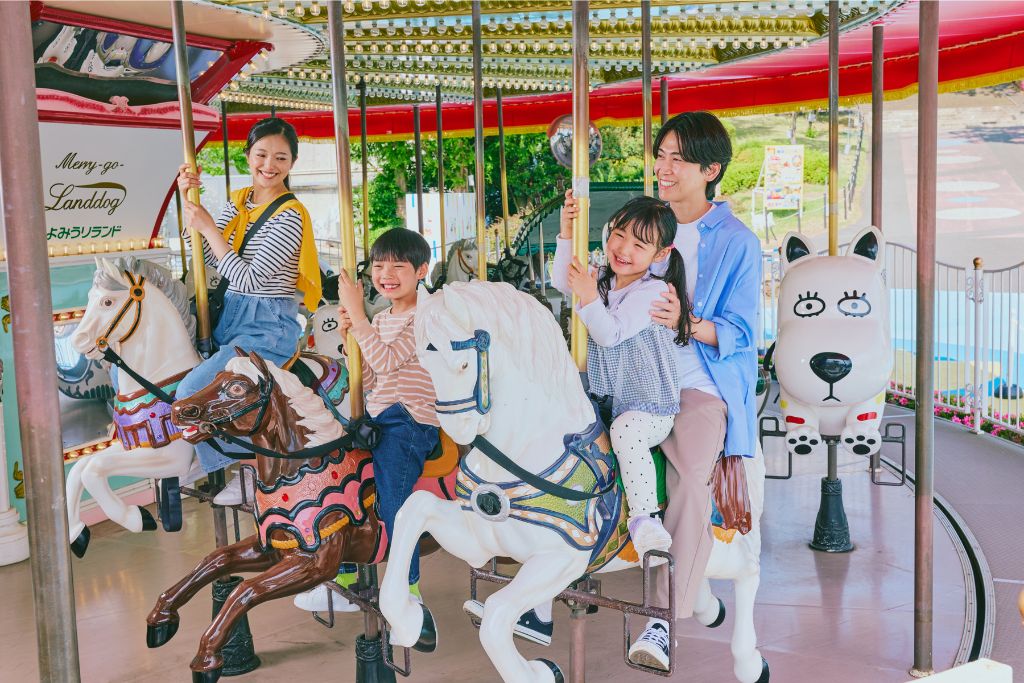 メリーゴーラウンドの白い馬と茶色い馬に乗る子どもと、支える大人。奥に犬の形をした乗り物が見える