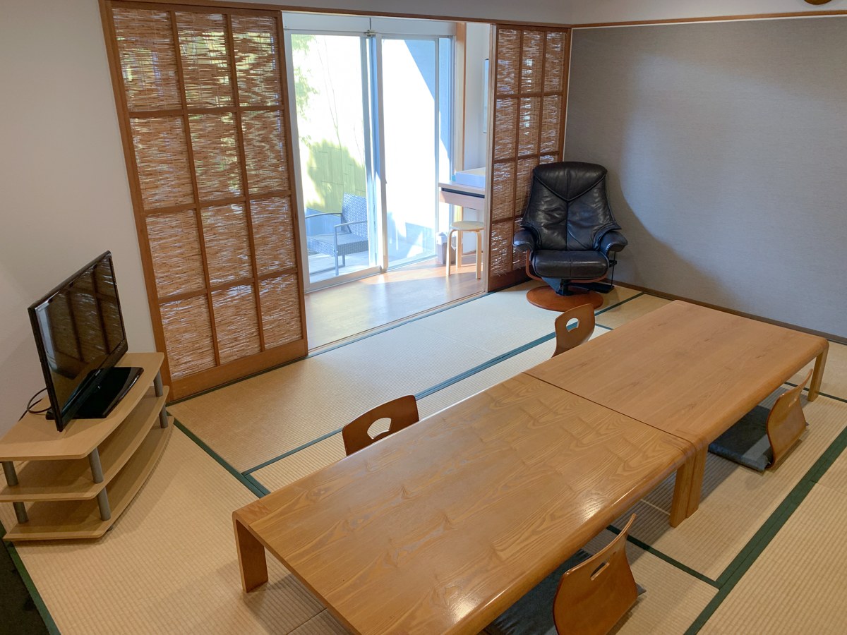 障子を開けた畳の部屋。座椅子とテレビ台が置かれ、窓の外に露天風呂が見える